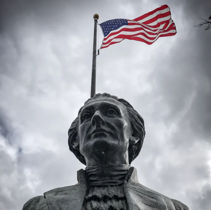 A bronze bust beneath an American flag under a cloudy sky